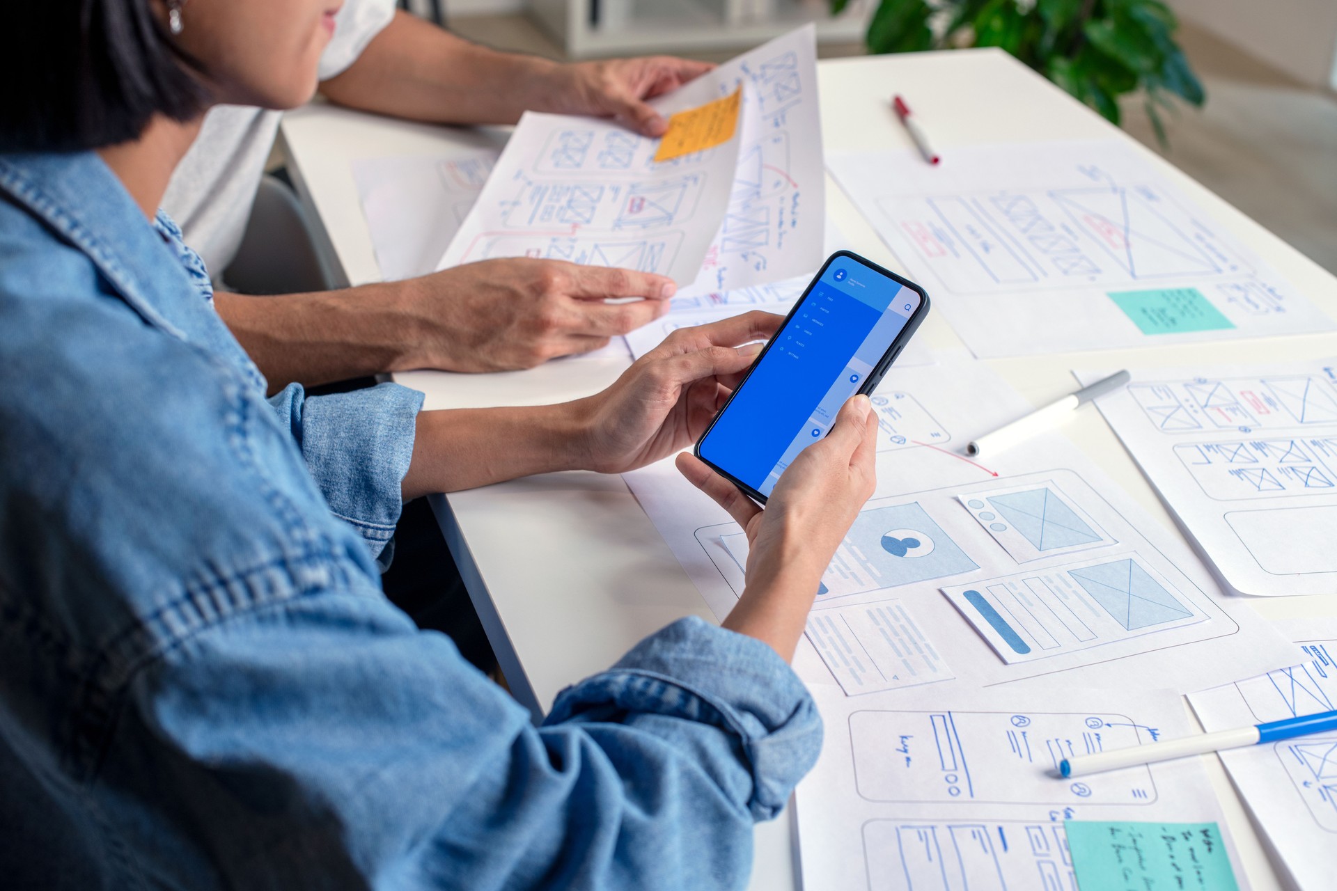 UX/UI designer testing prototype on a phone, discussing and brainstorming on wireframes for a website and mobile app prototype, surrounded by sketches of user flow and design tools, in the concept of website and mobile application design concept.